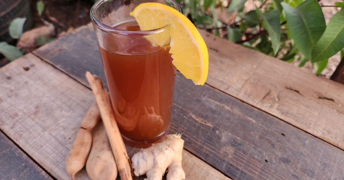 Close-up of Wassil Punch in glass with orange wedge on lip of glass with ginger, cinnamon and tamarind displayed in front on wood table outside.