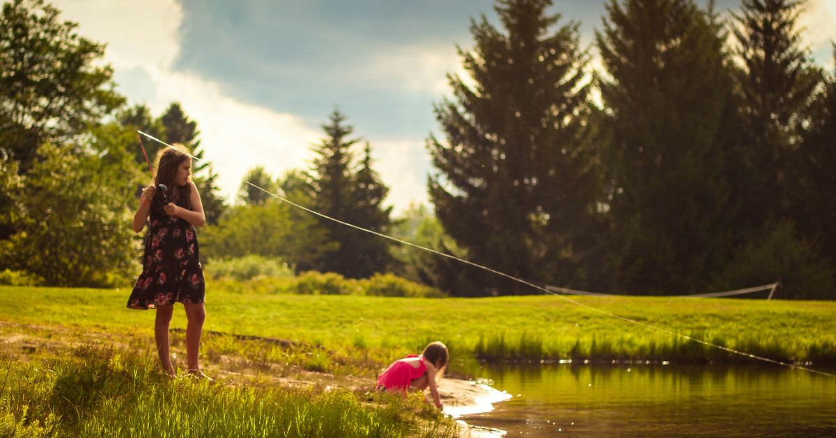 kid fishing on side of pond while another child playing at the side of pond.